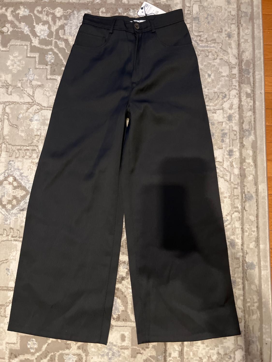 Black wide-leg pants with front pockets, button and zipper, placed on a geometric design rug in light tones.