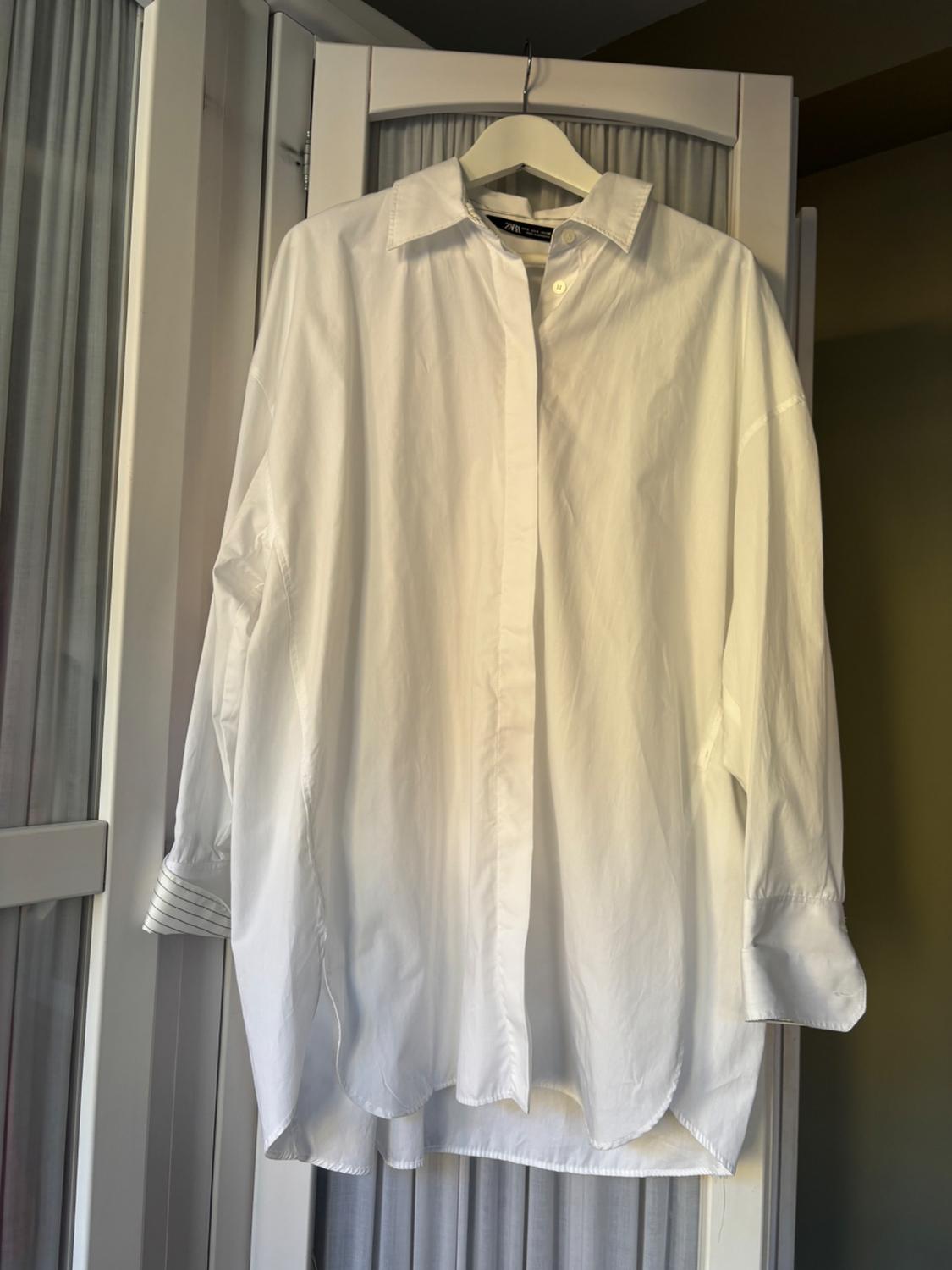 White, long-sleeved shirt hanging on a hanger in front of a white door with pleated curtains and warm light in the background.