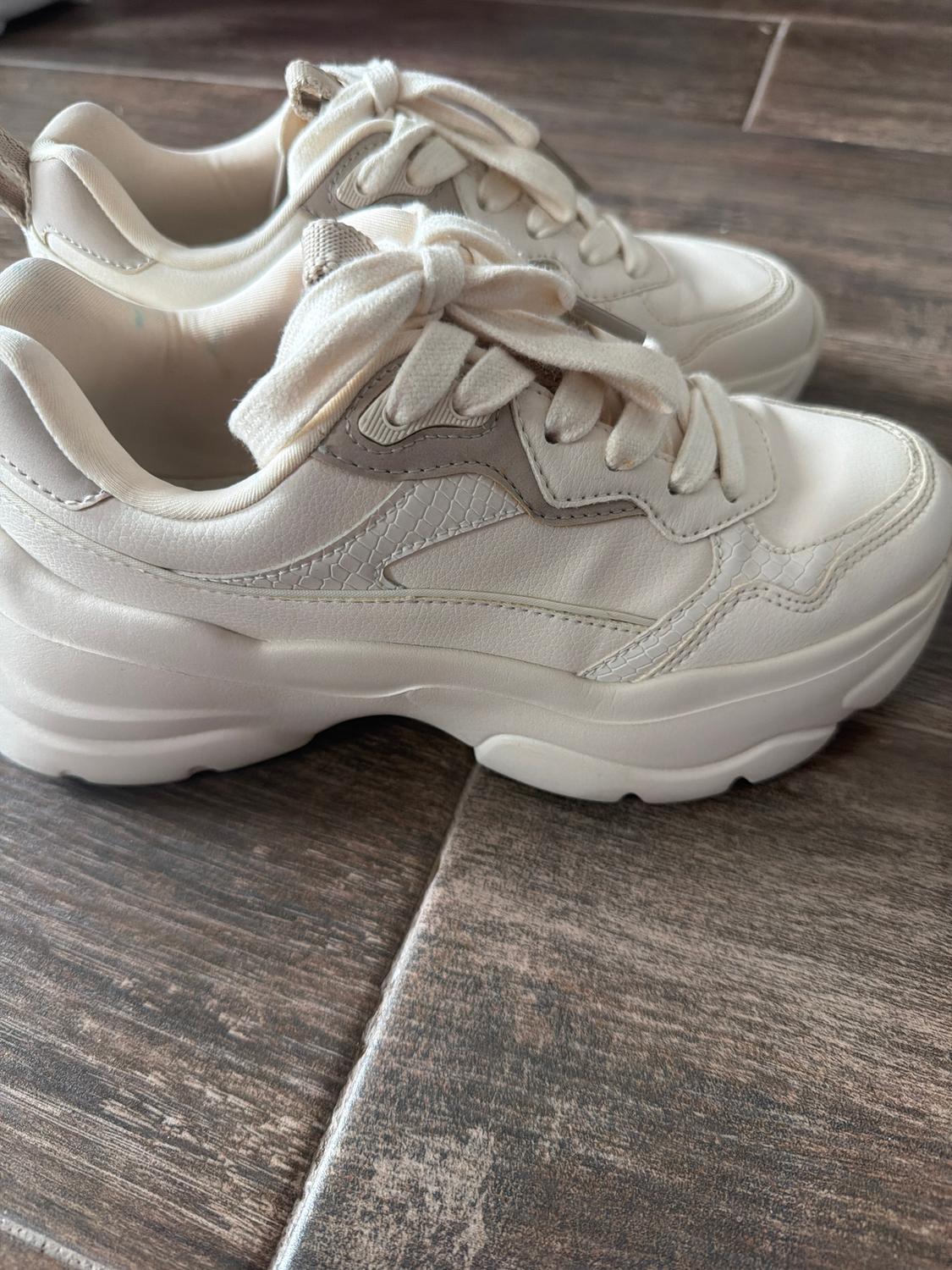 White trainers with a chunky sole and laces, on a dark wooden floor. Modern and casual design.