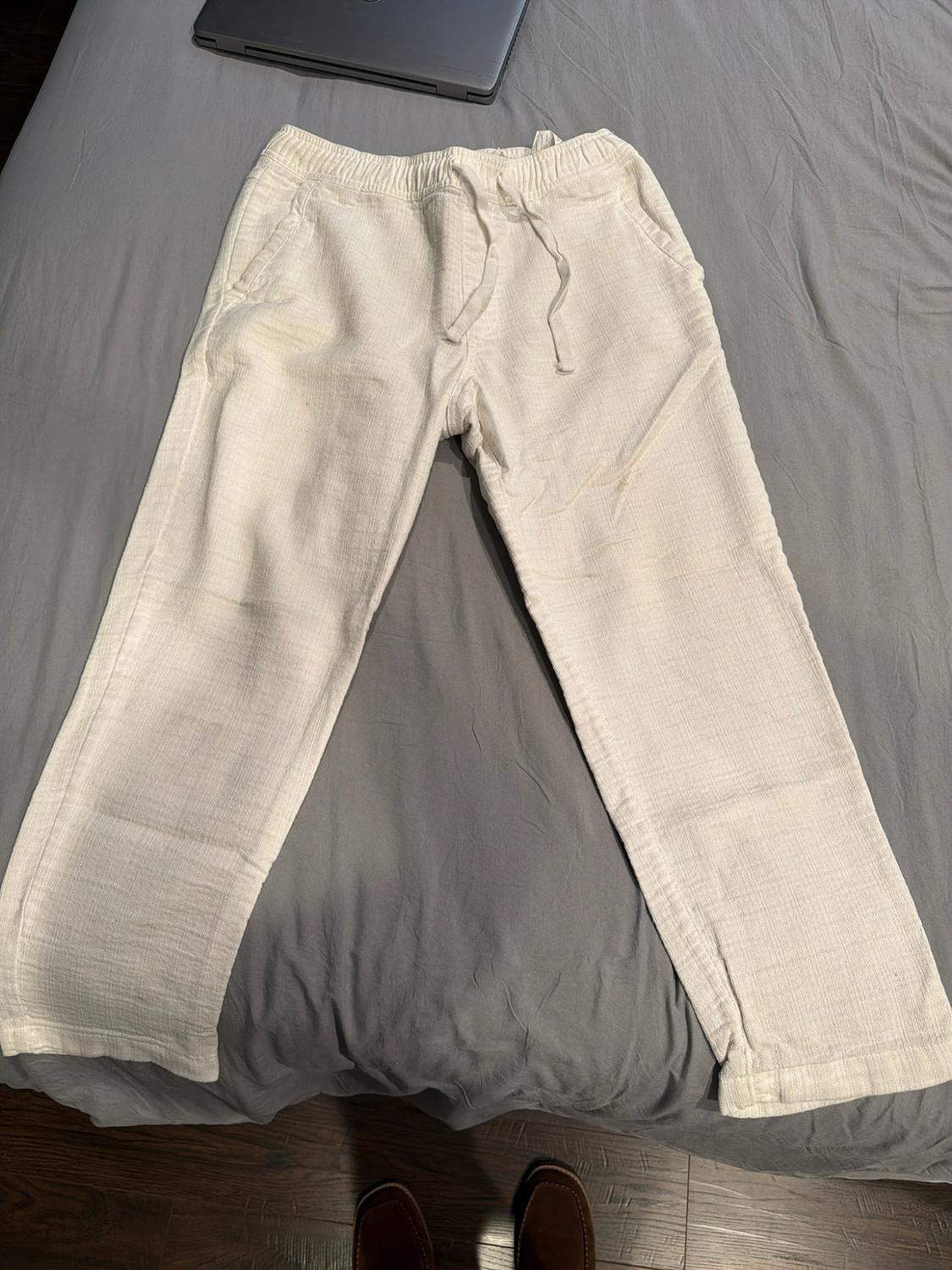 Casual white trousers with an elasticated drawstring waist, laid out on grey sheets. A laptop and shoes are visible. Relaxed atmosphere.