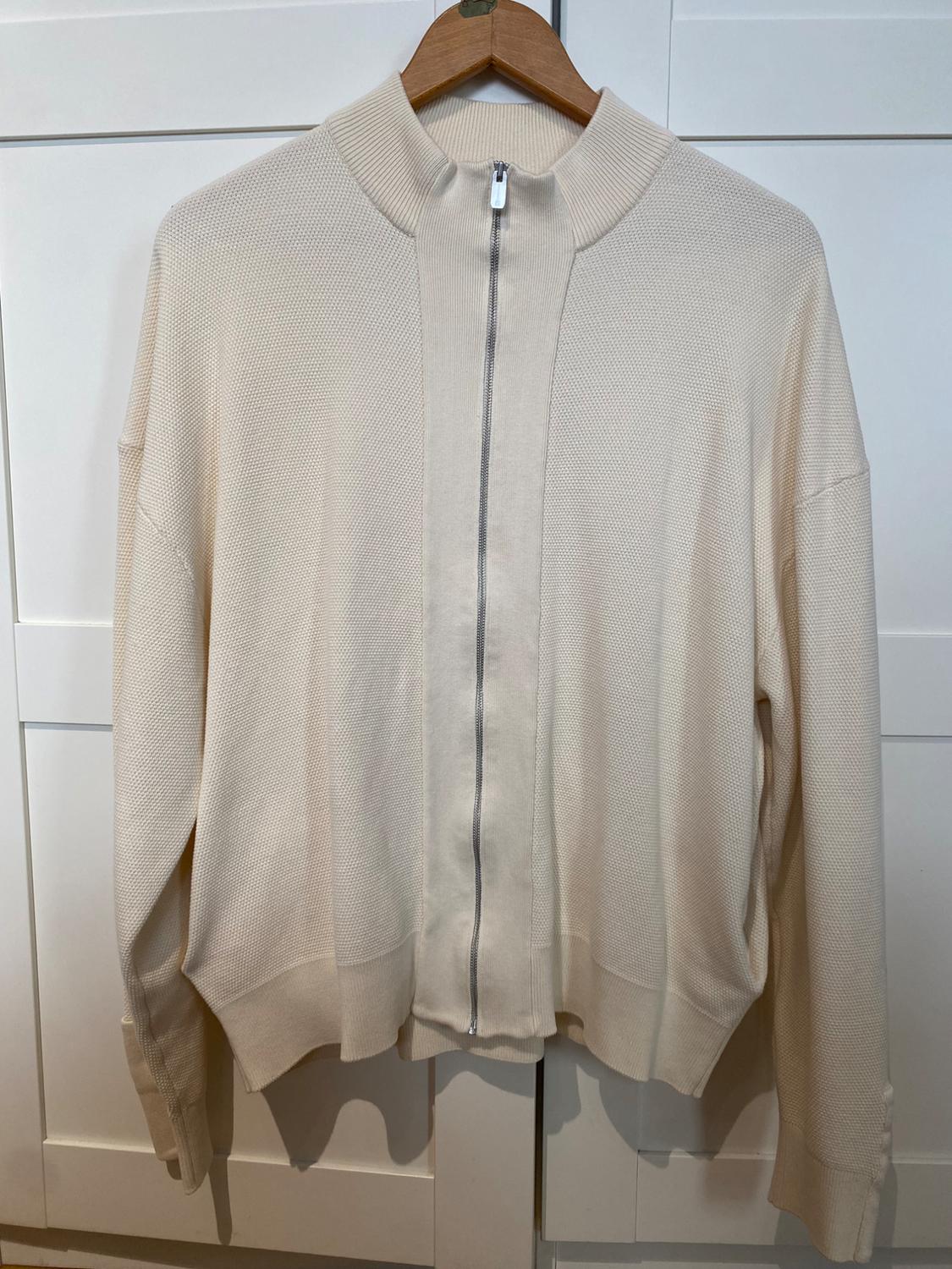 Beige knitted jacket with front zip, hanging on a wooden hanger in front of a white wardrobe.