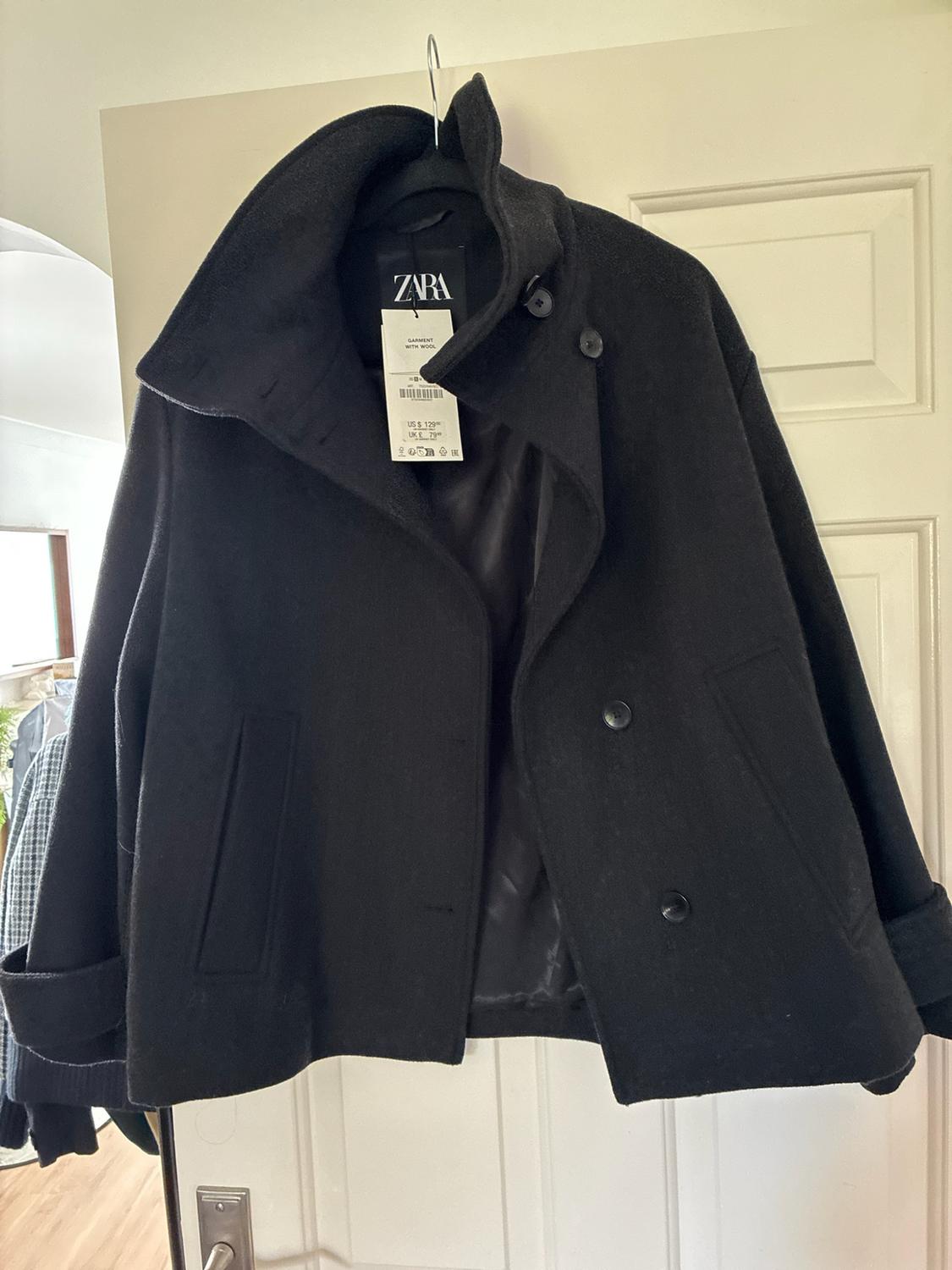Black high-neck buttoned coat hanging on a white door. Text: ZARA GARMENT WITH WOOL US $ 129.00 UK £ 79.00.