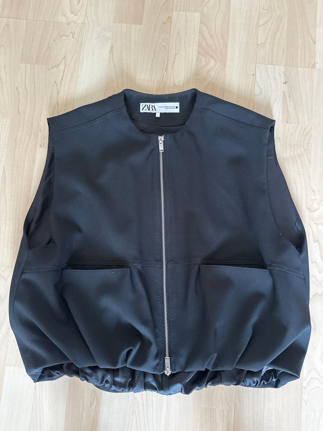 Black sleeveless gilet with front zip and pockets, on a light wooden floor. Visible label: "ZARA".