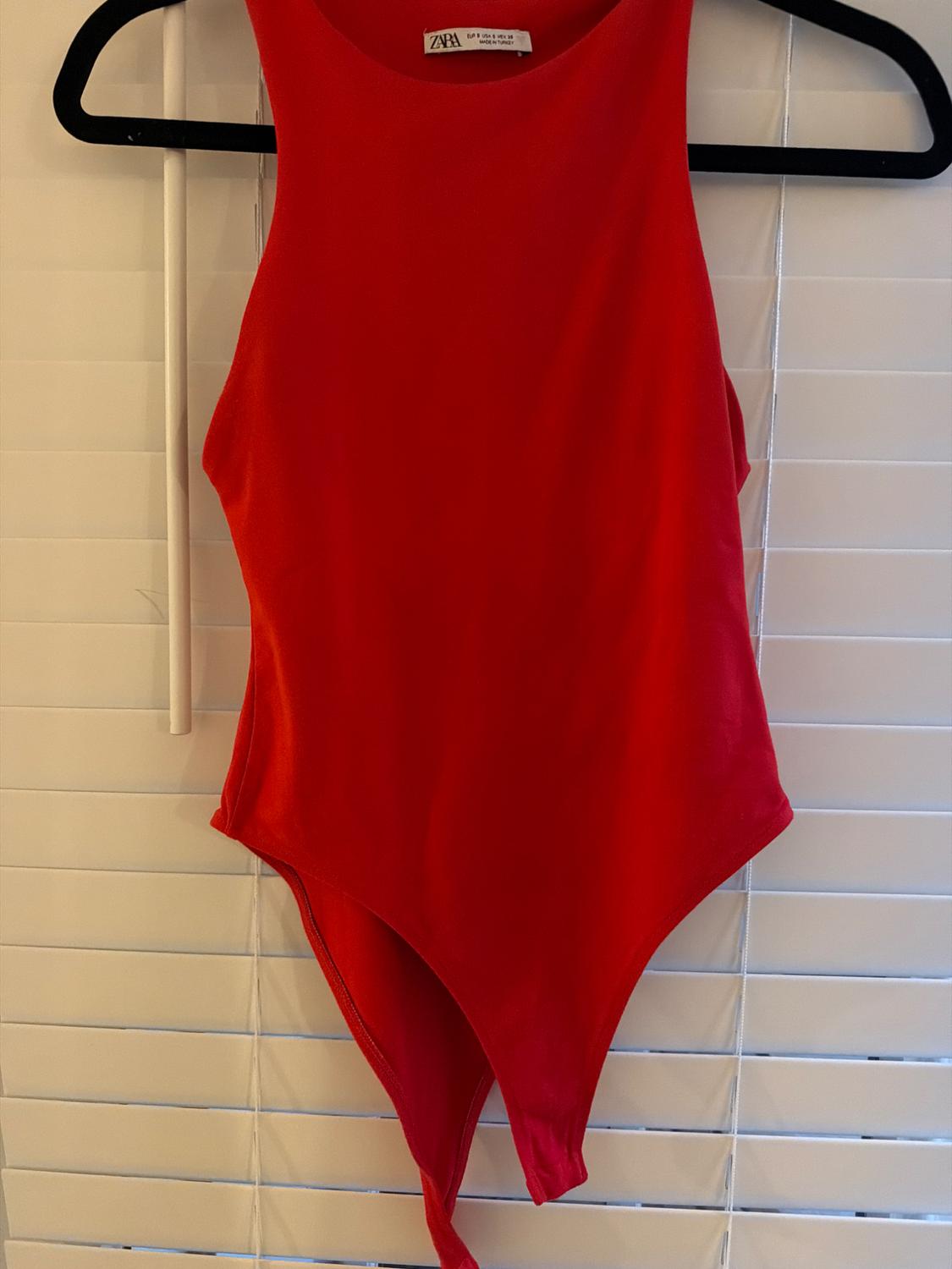 Red sleeveless bodysuit hanging on a black hanger in front of white blinds. Tag: ZARA EUR S USA S MEX 26 MADE IN TURKEY.