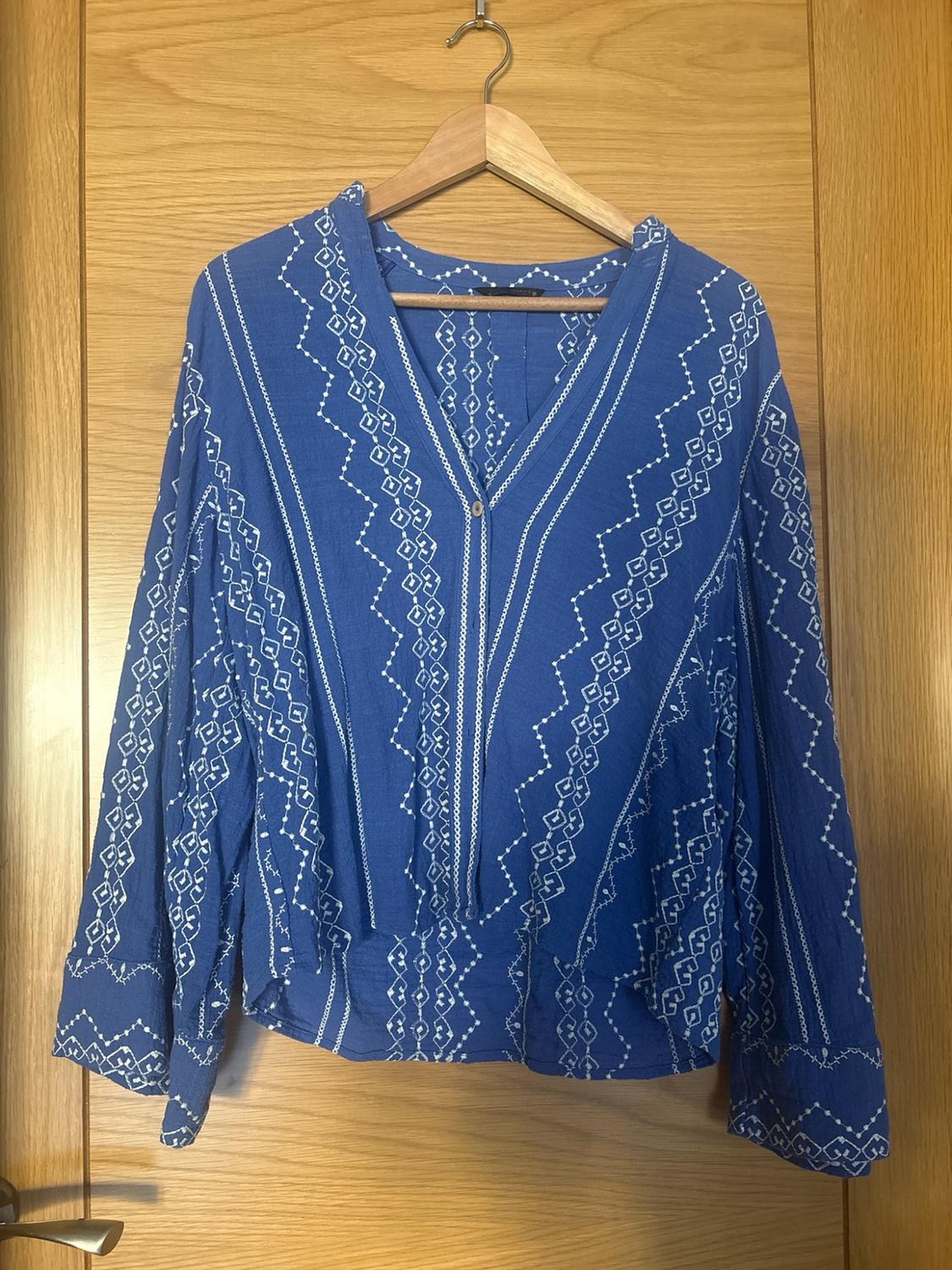 Blue long-sleeved V-neck blouse with white geometric patterns, hanging on a hanger on a wooden door.