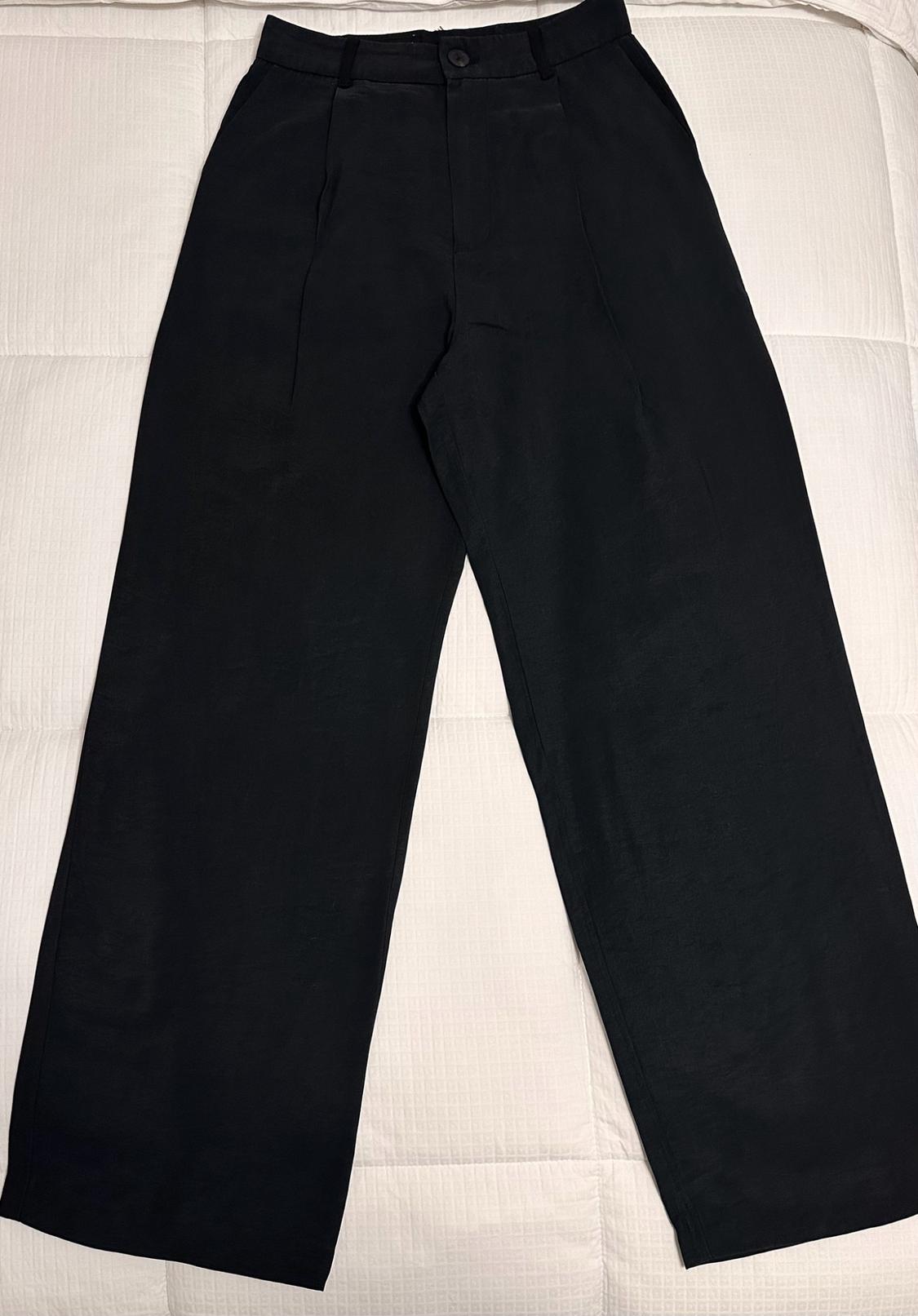 Black wide-leg pants with front pleats and a button closure, laid out on a padded white background.