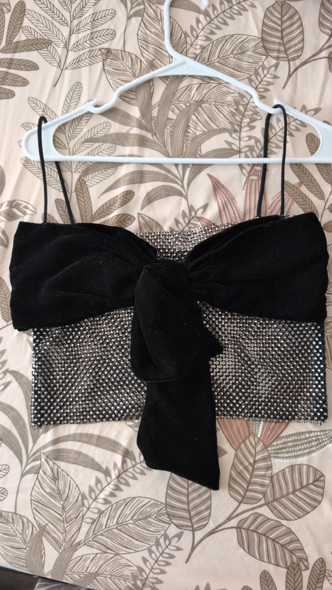 Thin strap camisole, rhinestone mesh and large black velvet bow, hanging on a hanger against a leaf-print background.