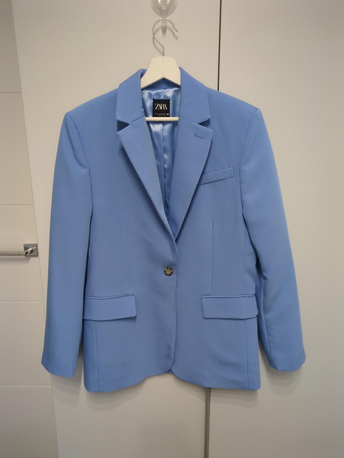 Ανοιχτό μπλε σακάκι Zara κρεμασμένο σε μια λευκή κρεμάστρα πάνω από μια λευκή πόρτα. Το σακάκι έχει ένα κουμπί και πέτα.