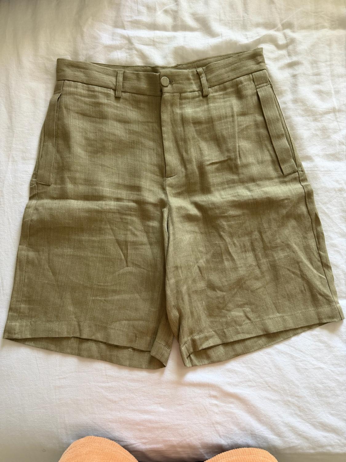 Light olive green shorts with pockets, laid out on a crumpled white sheet. A casual and relaxed style composition.
