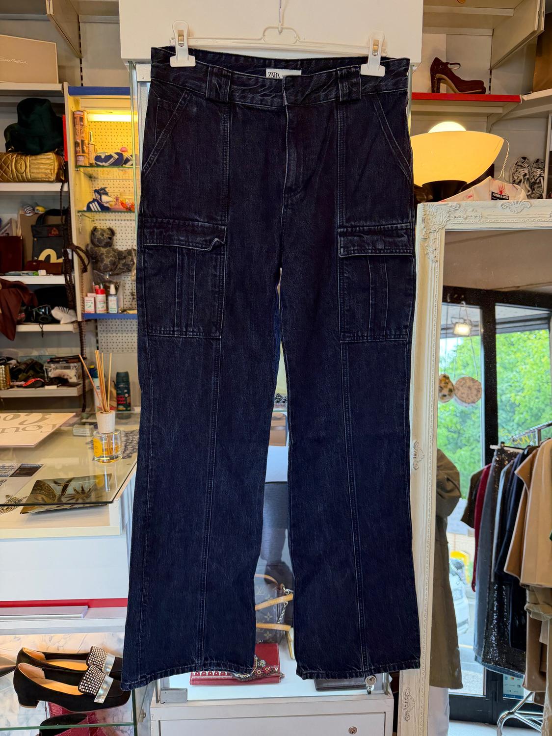 Dark cargo trousers hanging on a hanger in a shop, surrounded by clothes and accessories on shelves and in display cases.
