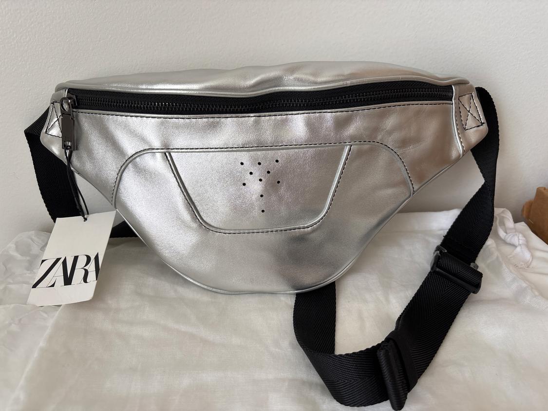 Silver fanny pack with black zipper and adjustable strap. Label with text "ZARA". White background.