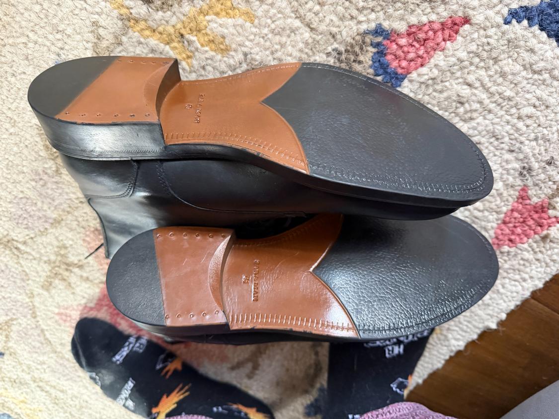 Black leather shoes with brown and black soles, on a colourful rug. Part of a foot with a dark sock visible.