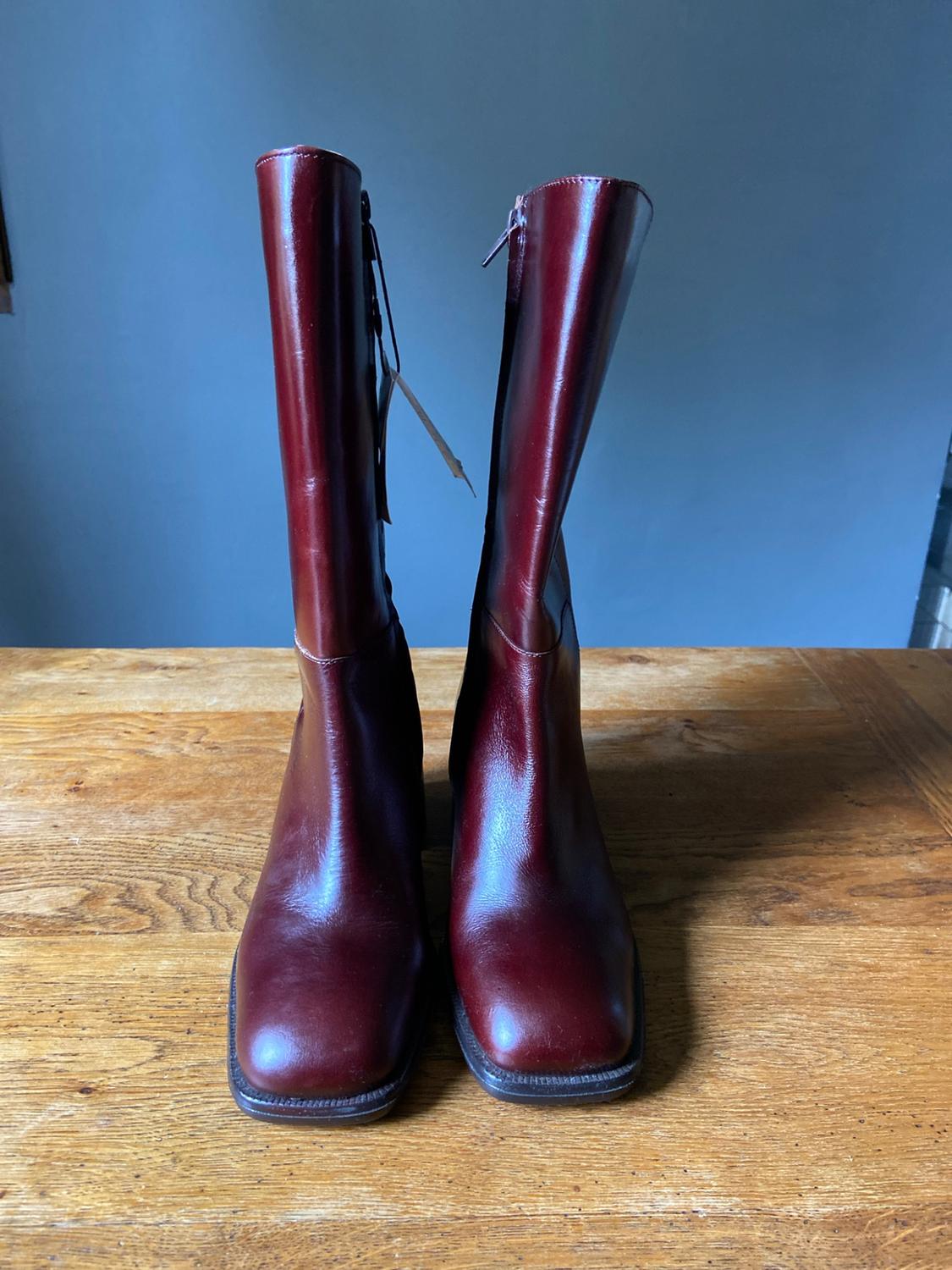 Burgundy leather knee-high boots with a square toe and zip fastening, resting on a wooden table in front of a blue wall.