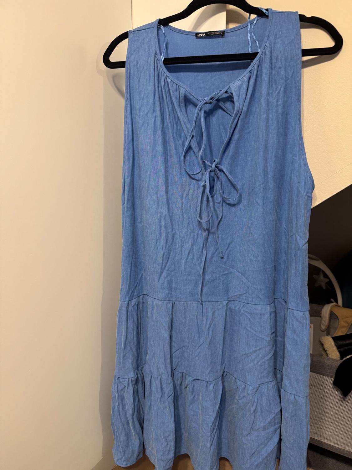 Sleeveless blue dress with a bow at the neckline, hanging on a black hanger. White wall and furniture background.