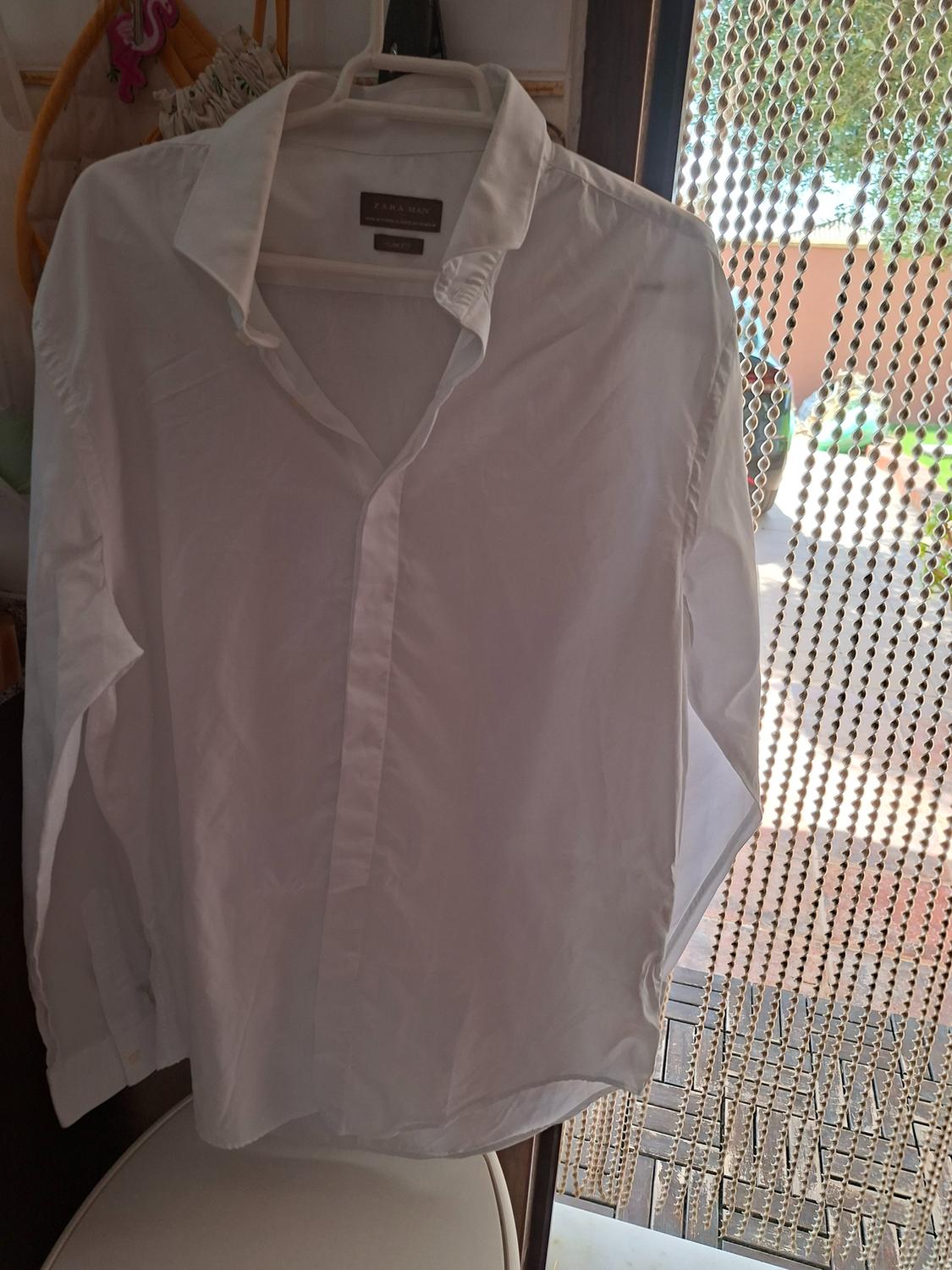 White long-sleeved shirt on a hanger. ZARA MAN label visible. Relaxed indoor setting next to a beaded curtain and natural light.