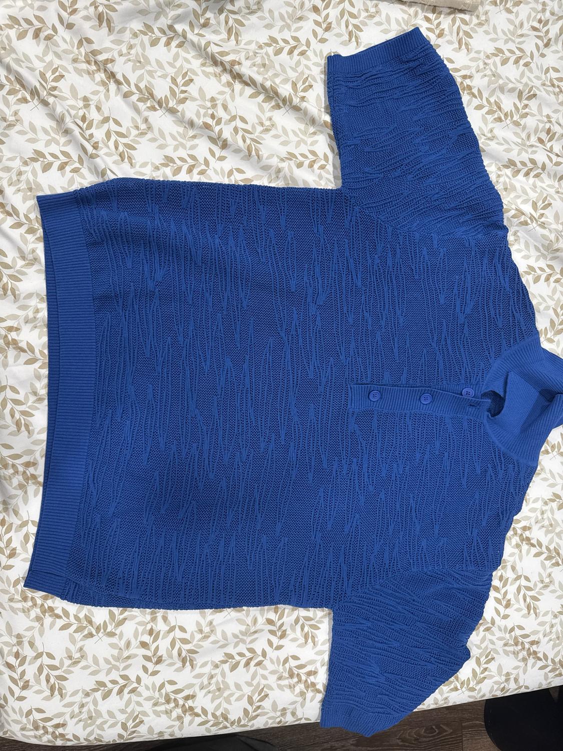 Blue knitted polo shirt with a wavy texture and short sleeves, laid on a white sheet with a beige leaf print. Casual style.
