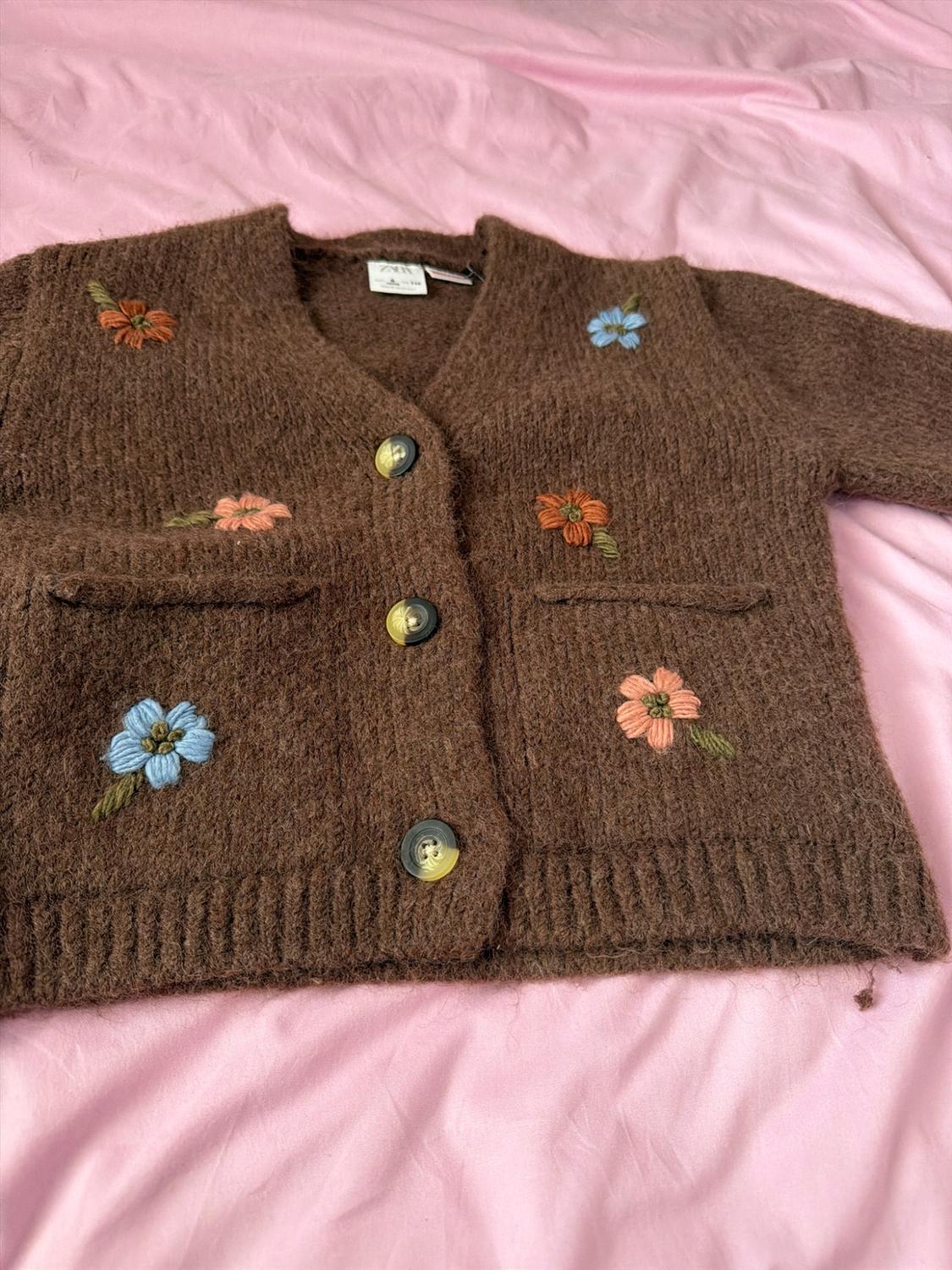 Brown knit cardigan with embroidered flowers, buttons and pockets, on a pink fabric. Visible ZARA label.