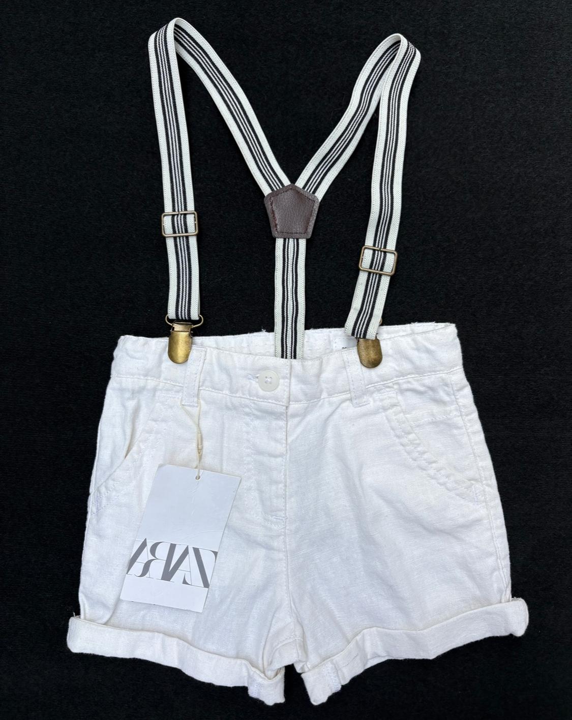 White shorts with black and white striped braces against a black background. Label with "ZARA" text hanging from the pocket.