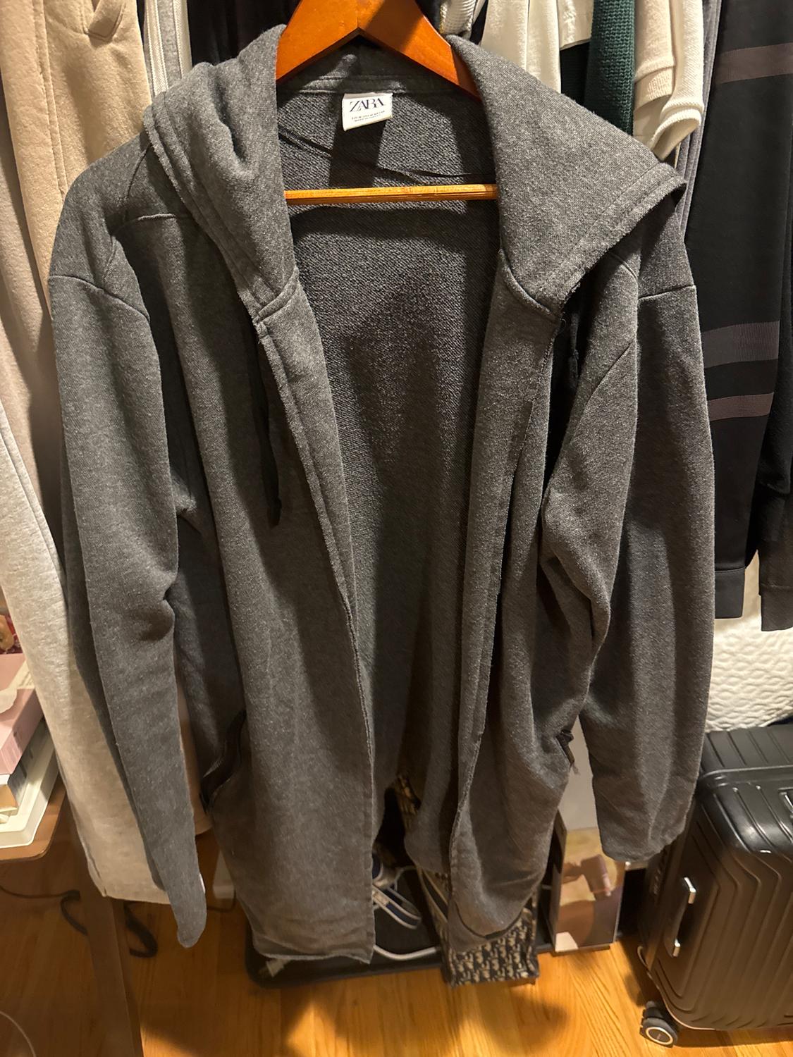 Grey hooded coat hanging on a wooden hanger in a wardrobe. Visible label with the text "ZARA". Background with clothes and suitcase.