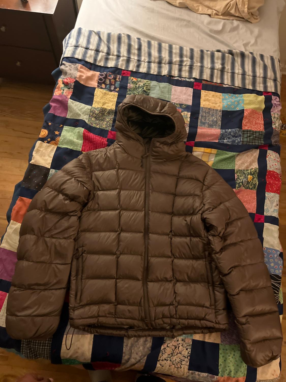 Brown padded jacket with a hood on a colourful patchwork quilt on a bed. Background with striped sheets.