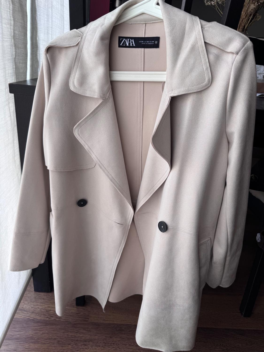 Women's beige coat hanging on a white hanger, with black buttons and a visible Zara label, against a wooden background and white curtain.