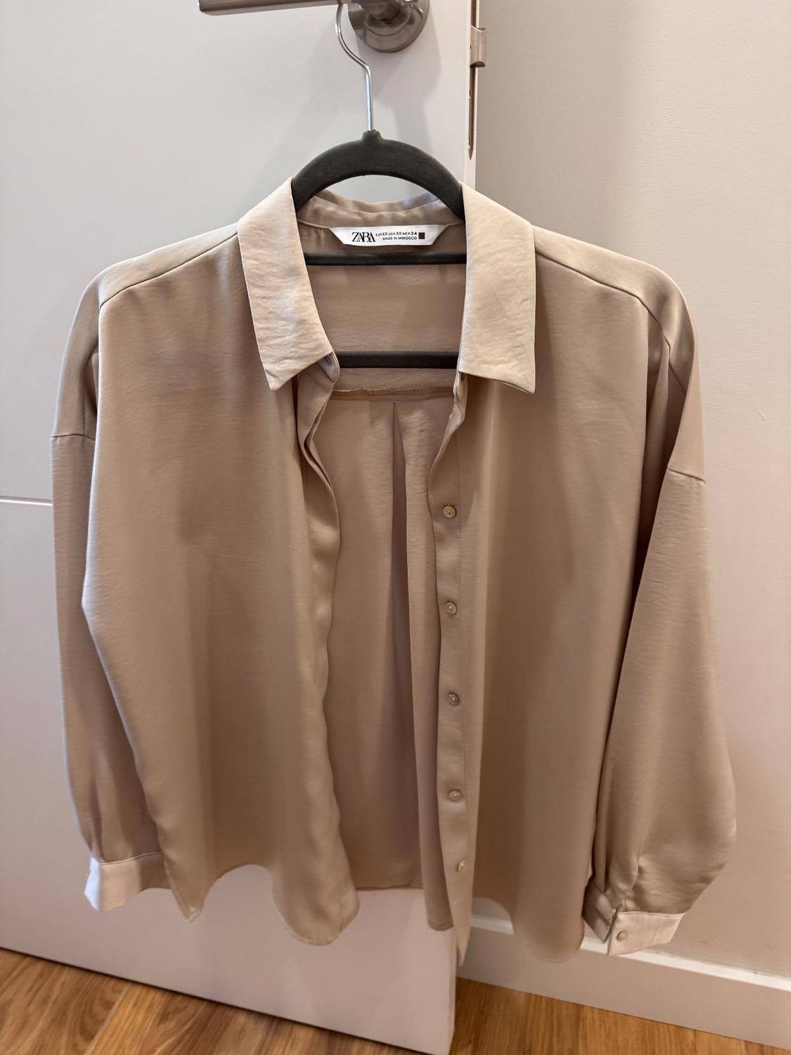 Beige long-sleeved shirt hanging on a black hanger over a white door. Visible label with text "ZARA".