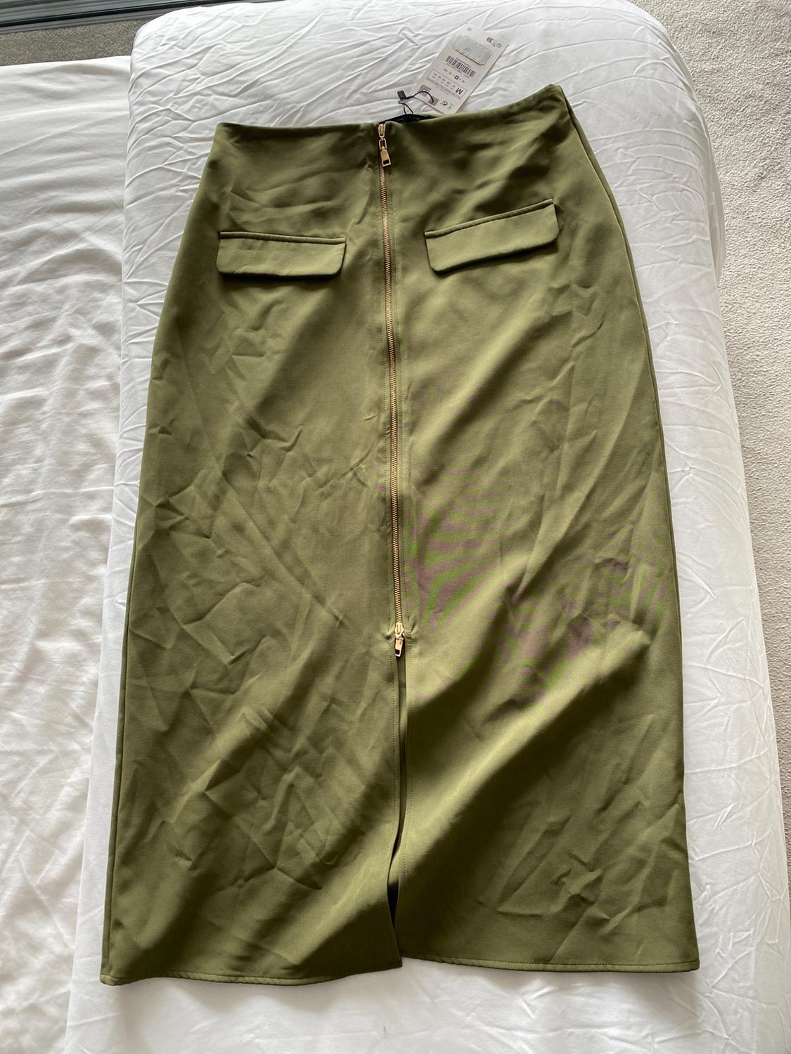 Olive green midi skirt with a central gold zip and two decorative pockets, placed on a bed with white sheets.
