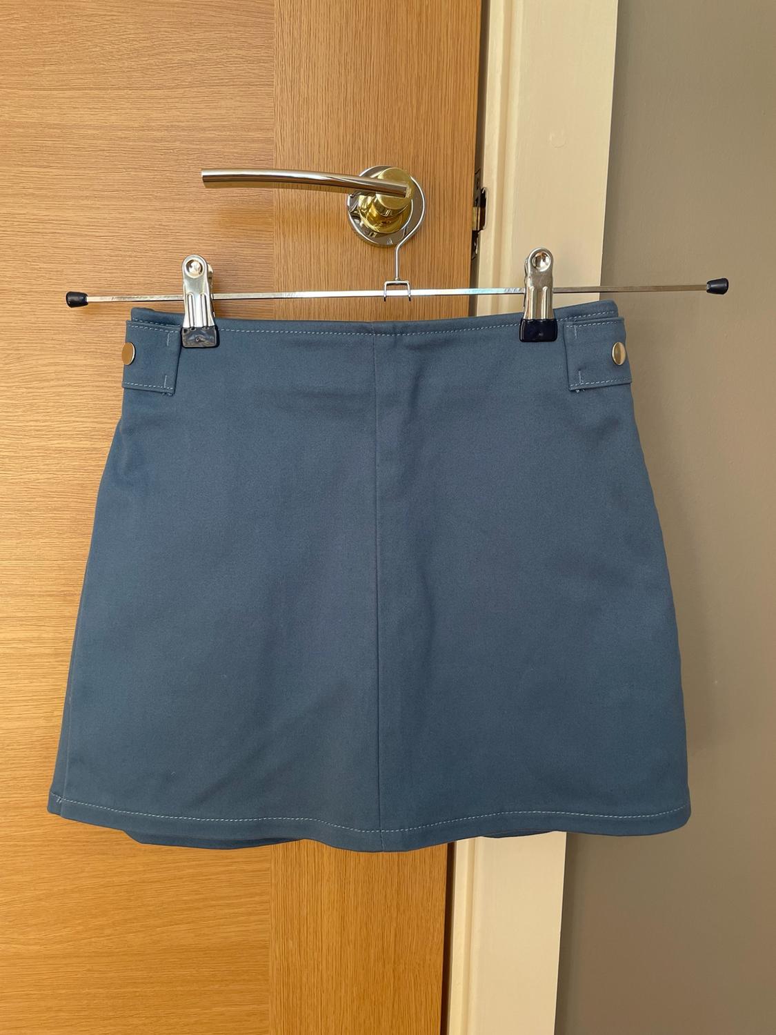 Blue miniskirt hanging from a hanger on a wooden door knob. It features visible stitching and button details on the waistband.