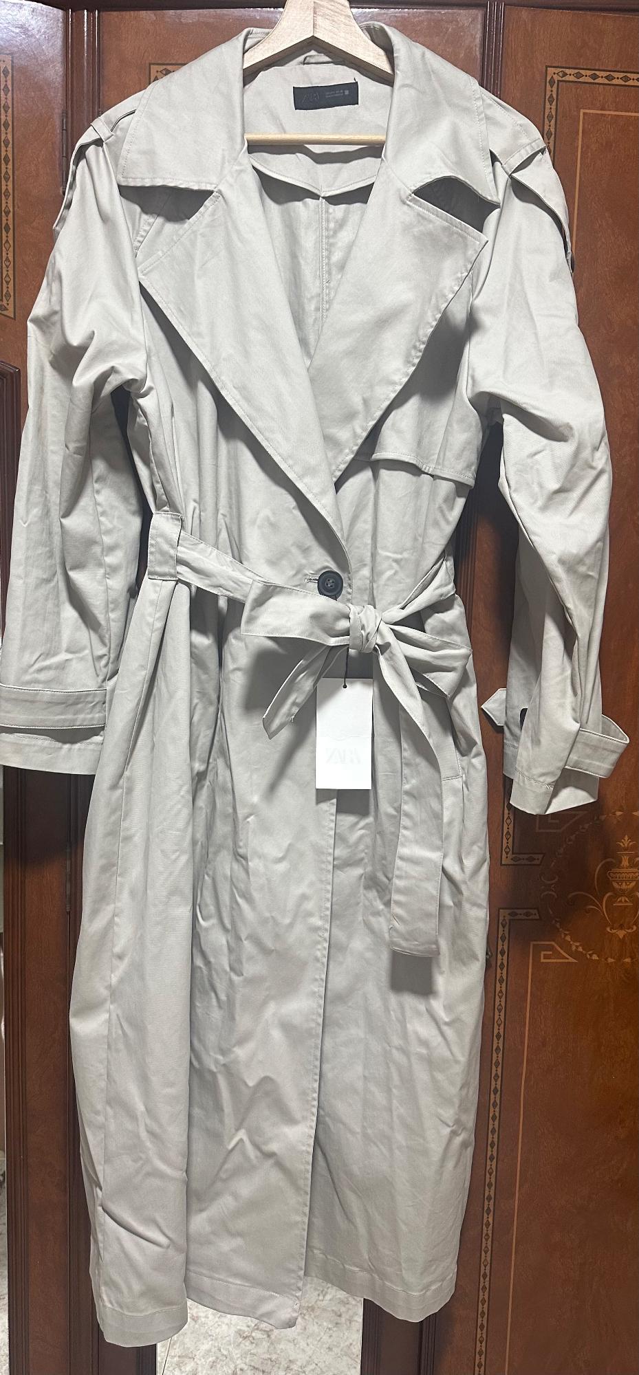 Beige trench coat hanging on a wooden hanger, with a tied belt and visible label. Background of decorated wooden doors.