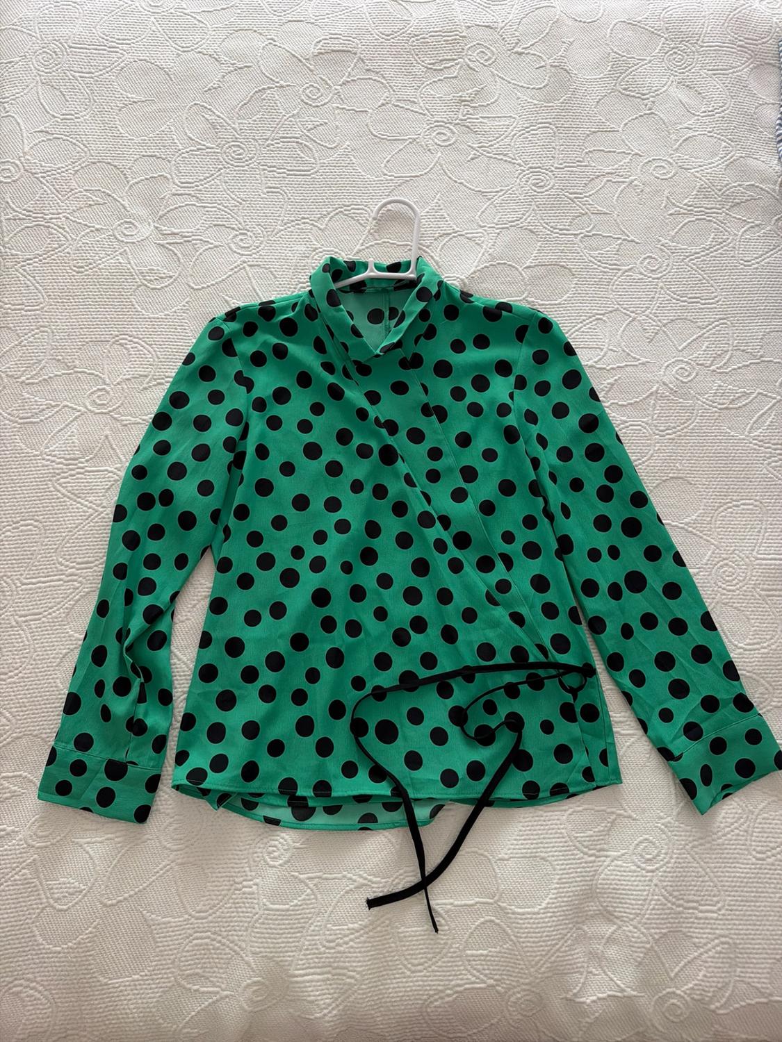 Green blouse with black polka dots, long sleeves and a high neck on a hanger, on a textured white bedspread with a casual and relaxed feel.