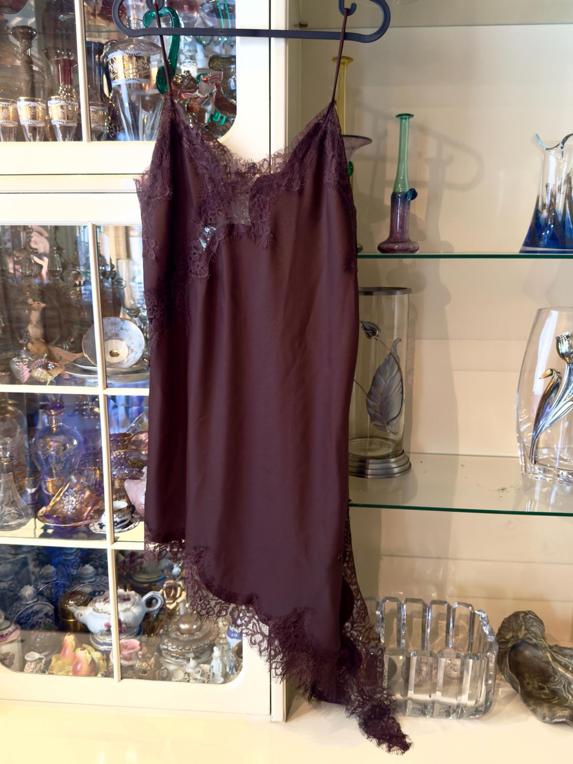 Burgundy slip dress with lace at the neckline and an asymmetrical hem, hanging on a hanger in front of a display cabinet with decorative glassware.