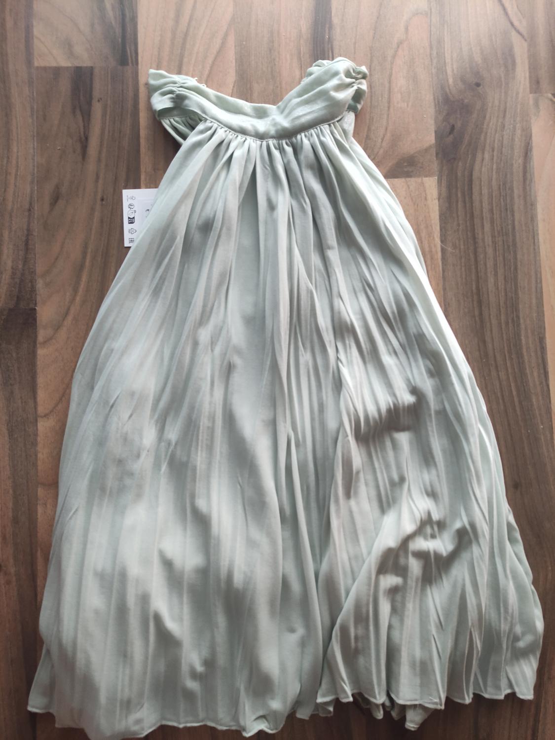 Long pleated dress in a light green tone against a wooden backdrop. Label visible to the left of the dress.
