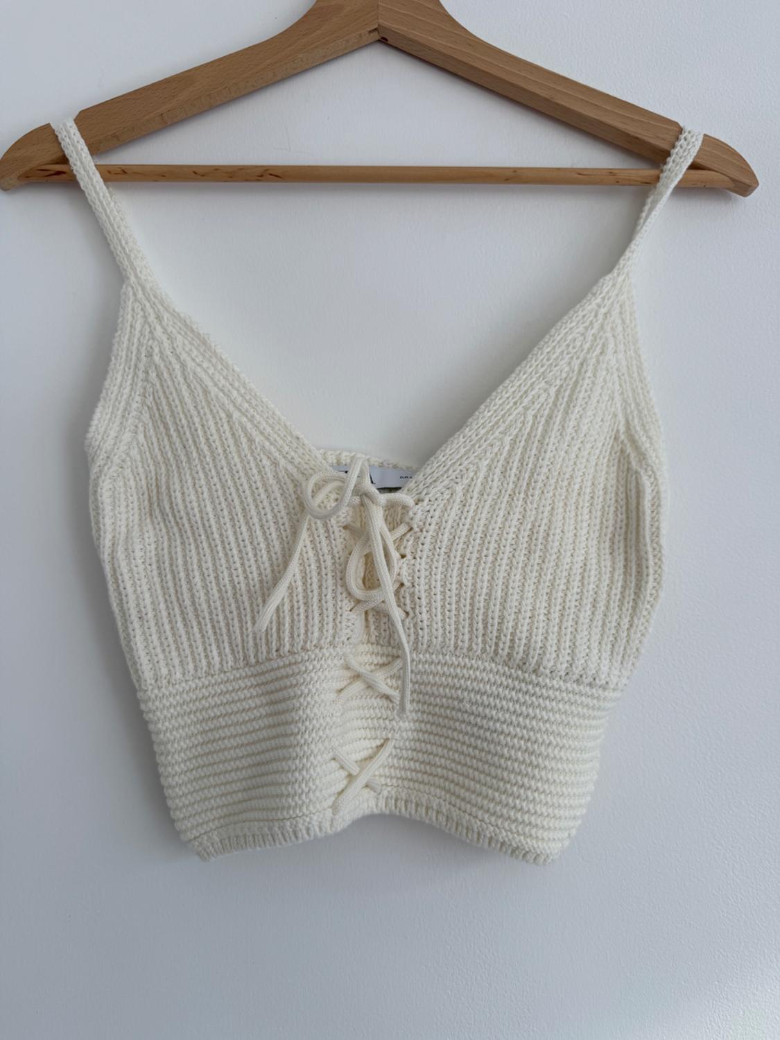White knitted top with spaghetti straps and a V-neck, hanging on a wooden hanger against a white wall.