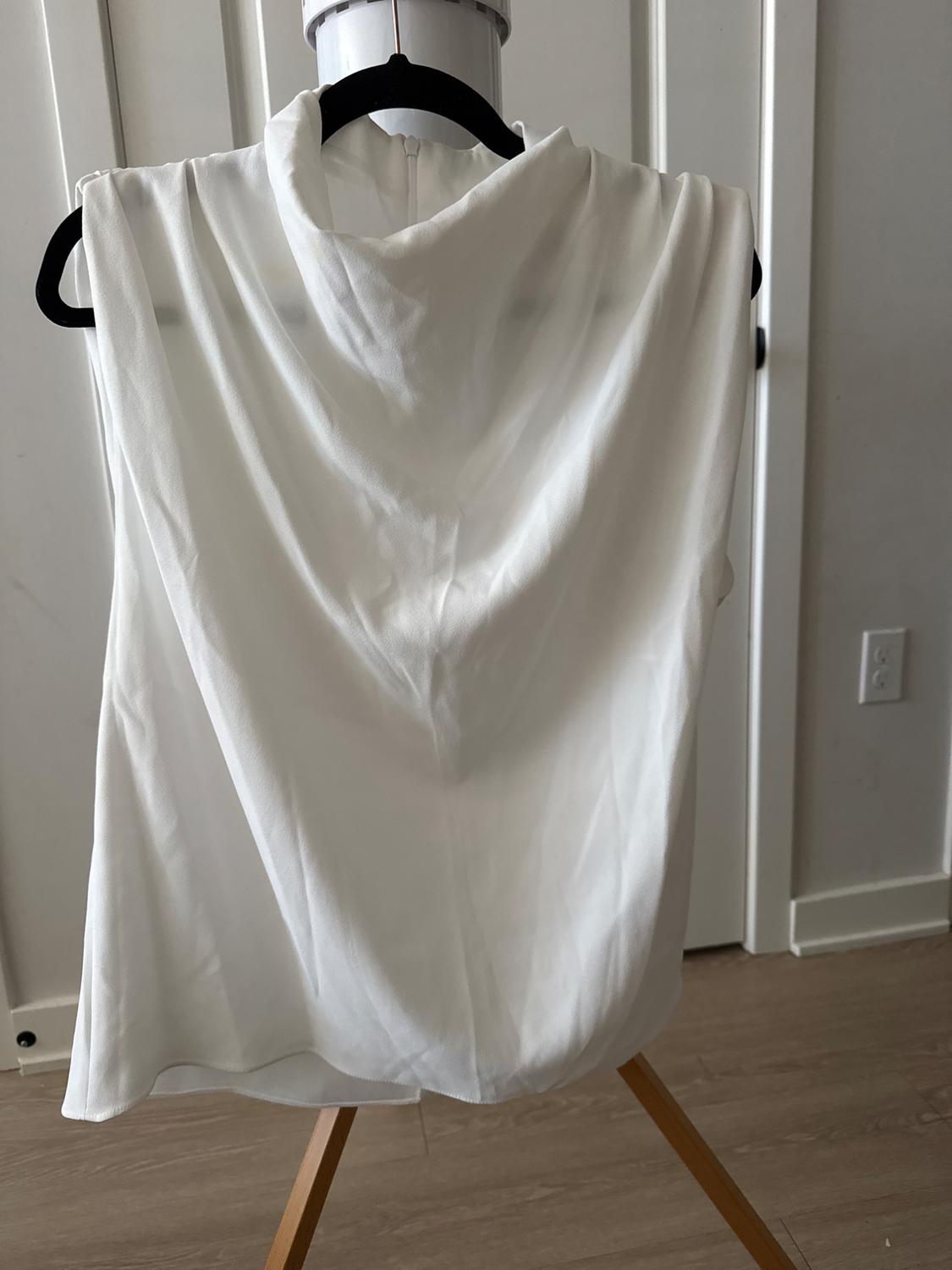 White, sleeveless blouse with a cowl neck, displayed on a mannequin against a backdrop of a white wall and light wood floor.