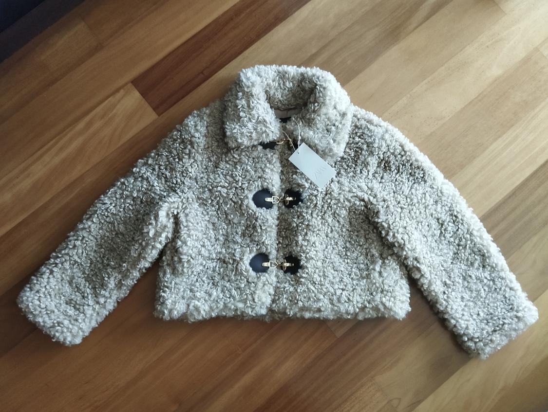 Beige teddy jacket with dark buttons on a light wooden floor. White label hanging from the collar.