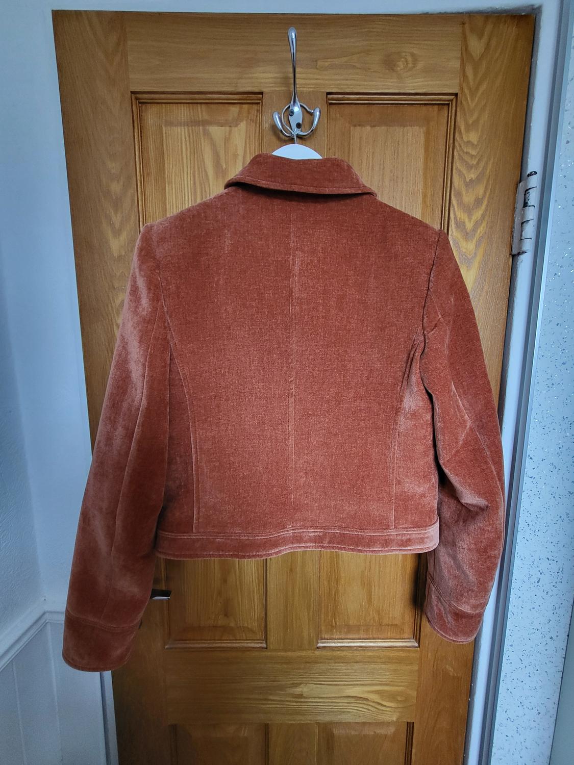 Terracotta velvet jacket seen from the back, hanging on a hook on a wooden door.
