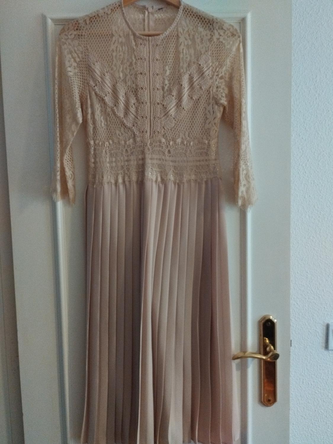 Beige lace dress with long sleeves and pleated skirt, hanging on a white door with a gold doorknob.