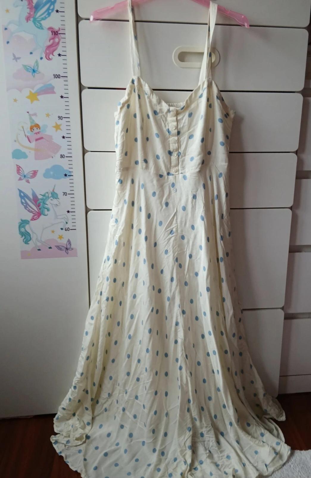 Long strap dress with blue polka dot print on a white background, hanging on a pink hanger in front of a white cabinet.