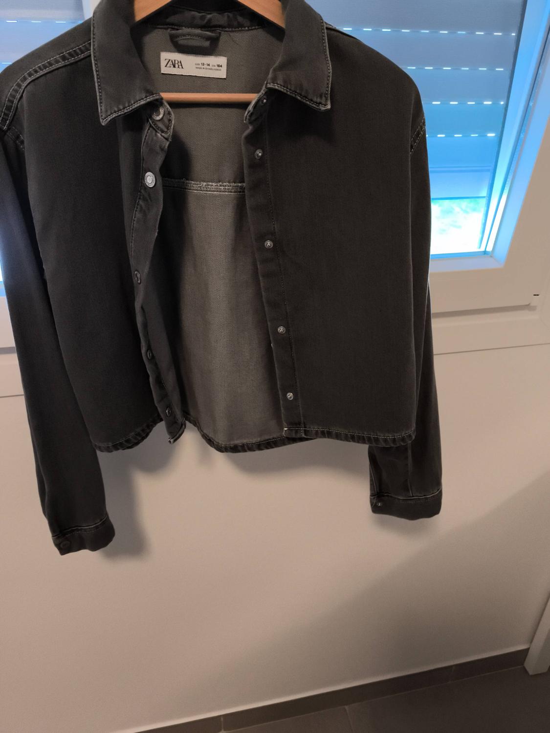 Black denim shirt hanging on a hanger in front of a window. Visible label with the text "ZARA".
