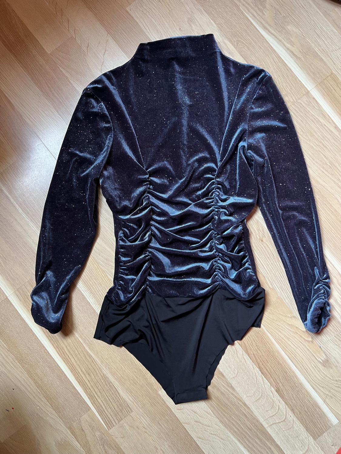 Black velvet bodysuit with long sleeves and ruching on the back, on a light wooden floor.