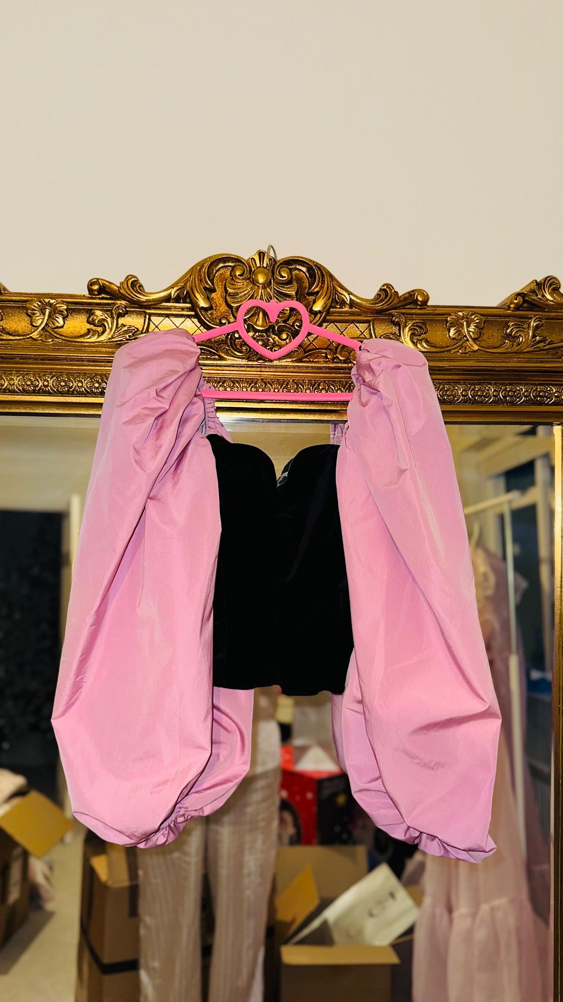 Black top with pink puff sleeves hanging on a pink heart hanger, reflected in an ornate gold-framed mirror.