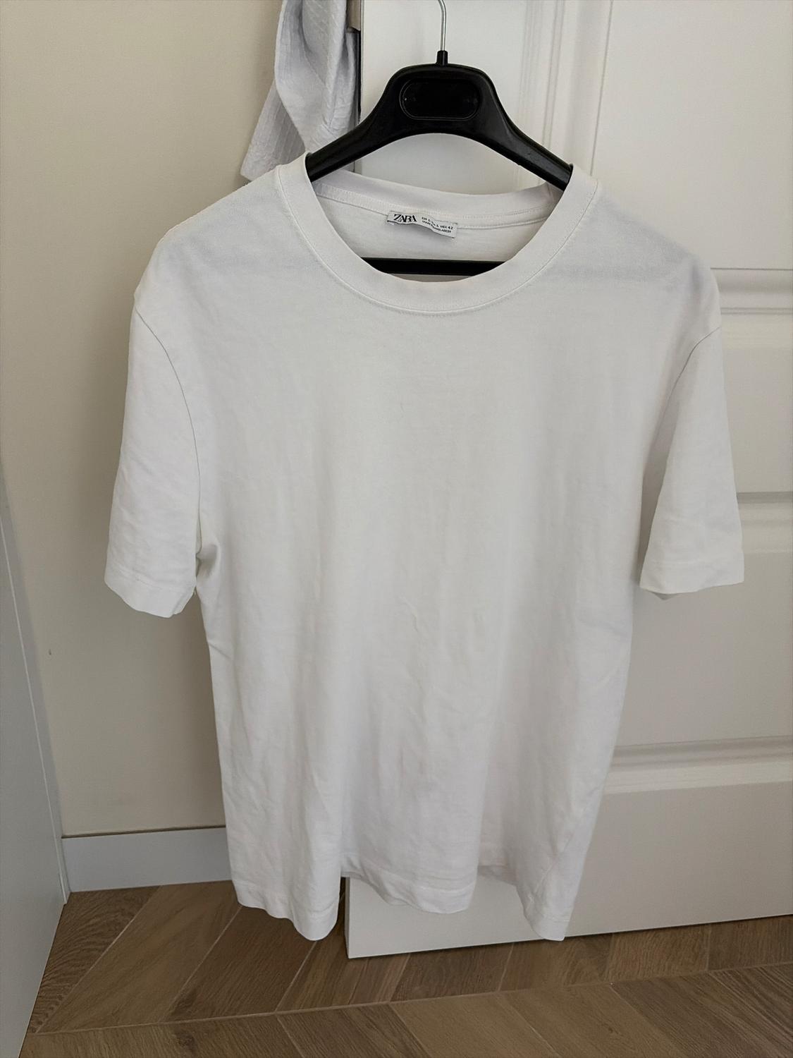 White short-sleeved t-shirt hanging on a black hanger in front of a white door. Wooden floor visible.