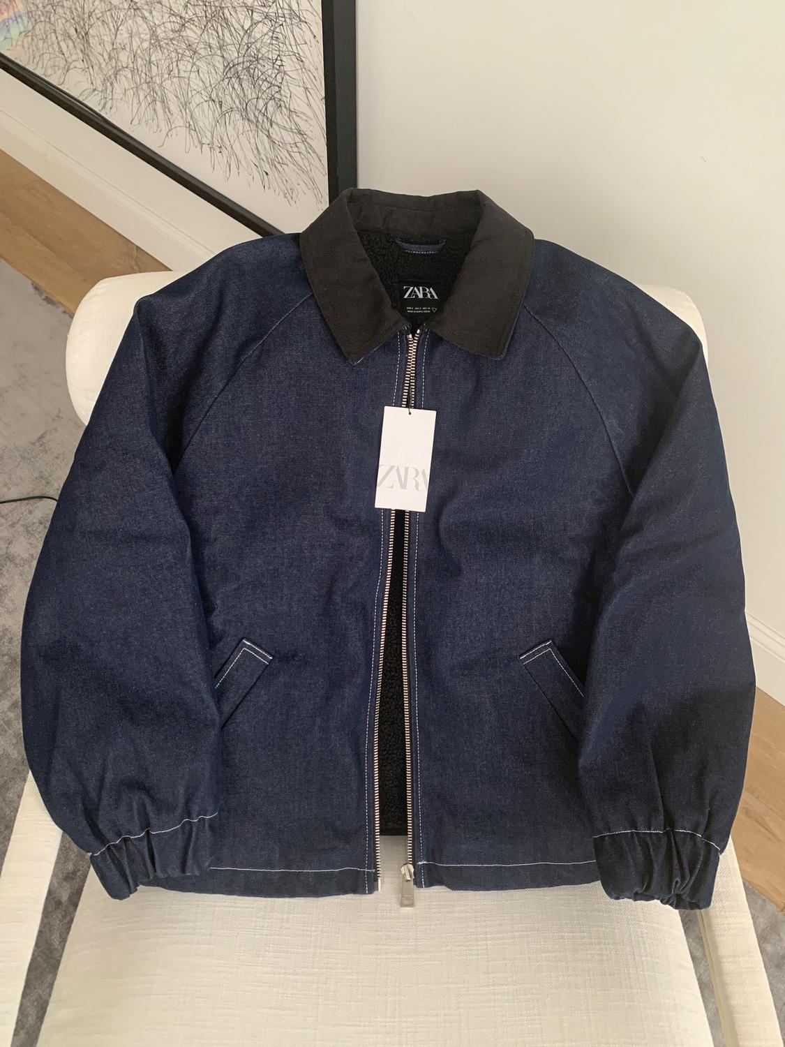 Blue zip-up jacket with black collar on white chair. Visible label with the text "ZARA". Background with white wall and line art.