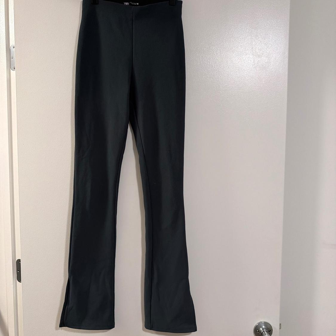 Dark-coloured, high-waisted, slim-fit trousers with flared hems and side slits, hanging on a hanger against a white door.