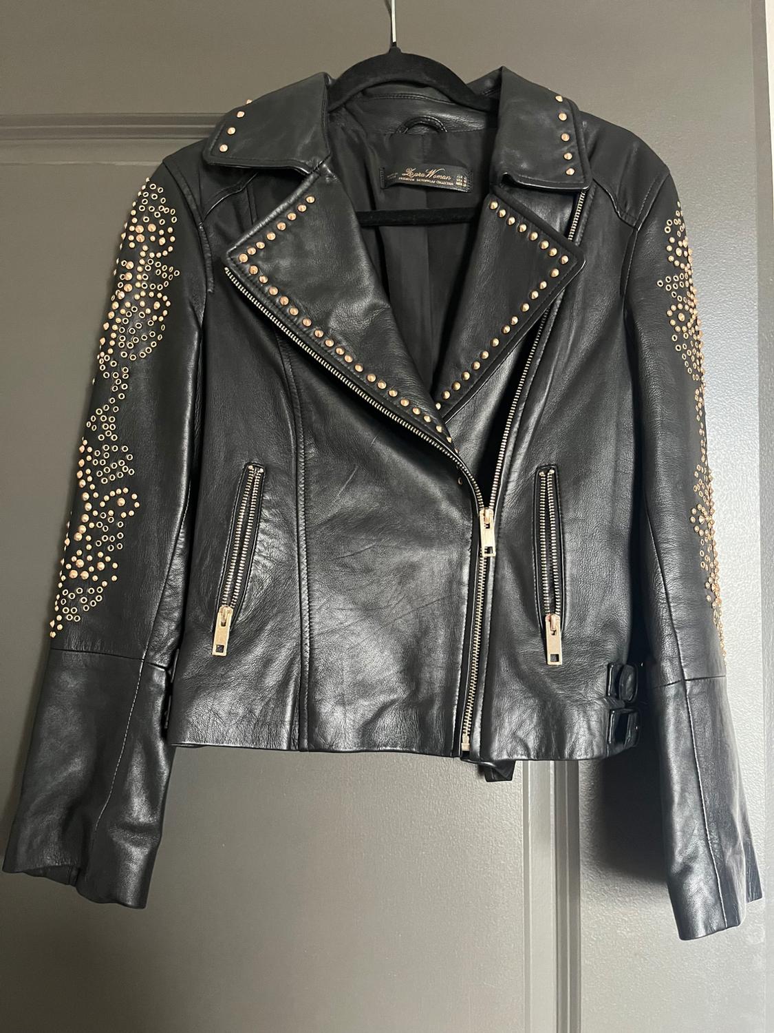 Black leather biker-style jacket with gold studs on the sleeves and lapels, hanging on a hanger over a gray door. Label: Zara Woman.