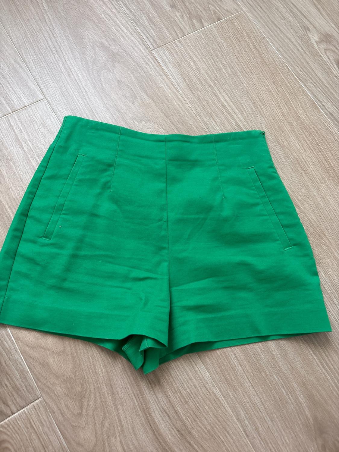 High-waisted green shorts with front pockets, laid out on a light wood floor in a relaxed flat lay composition.
