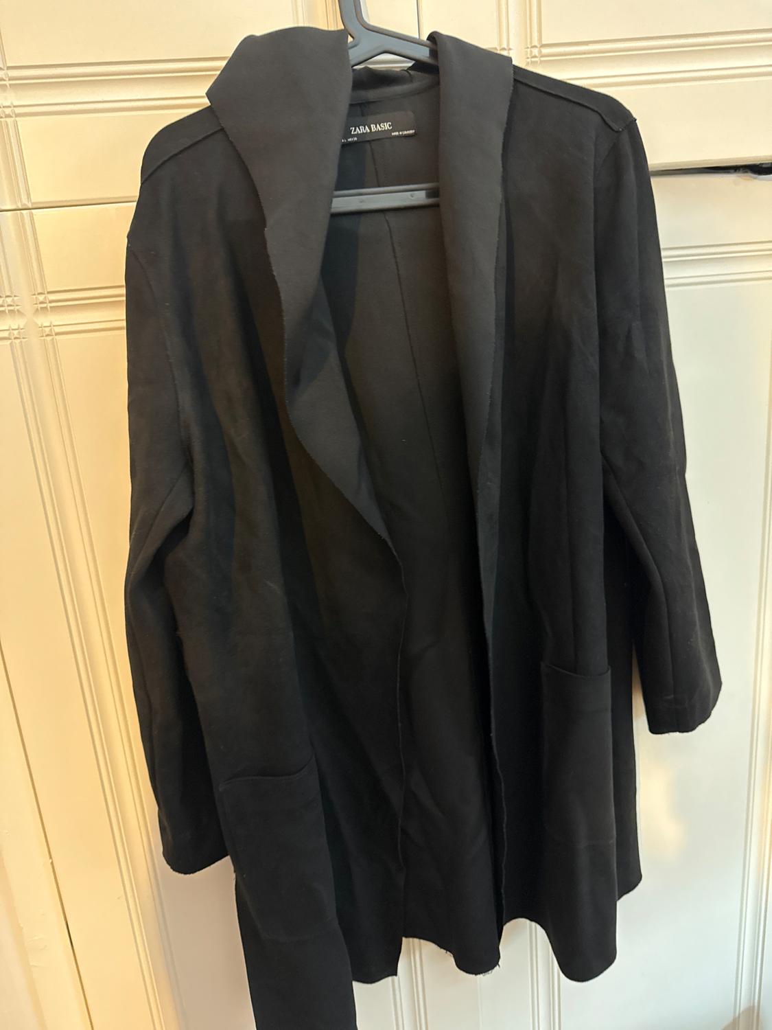 Black coat hanging on a hanger, with "ZARA BASIC" label. Light wood panel background.