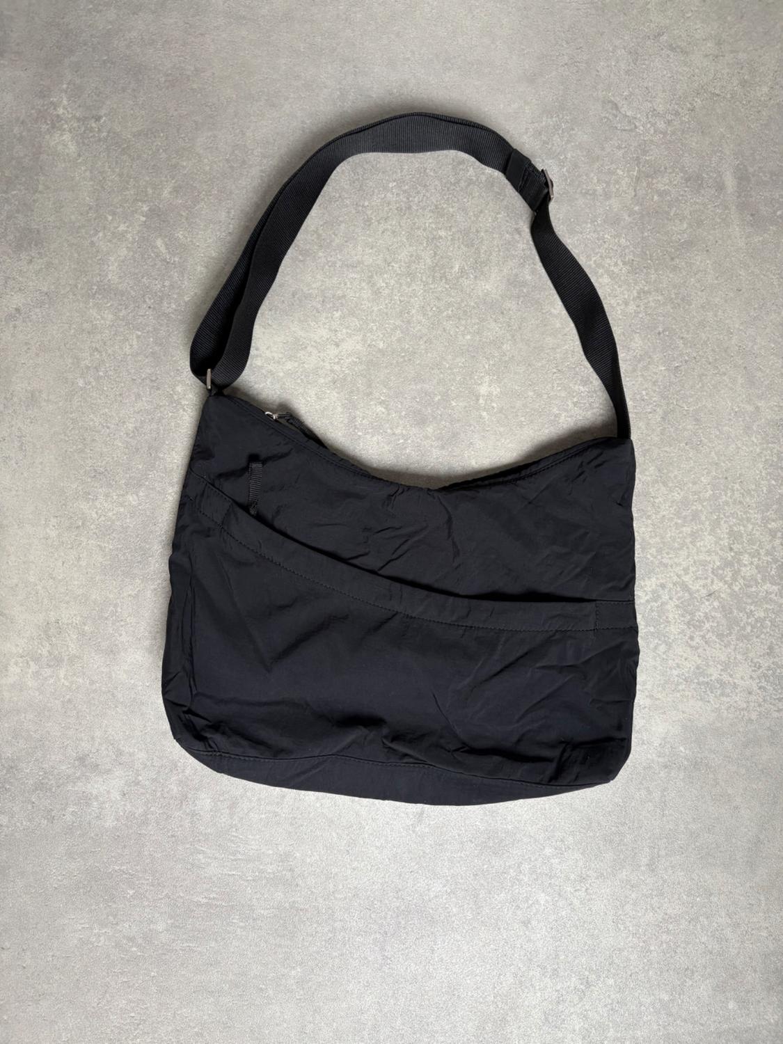 Minimalist black cross-body bag with an adjustable strap and front pocket detail, on a textured grey background.