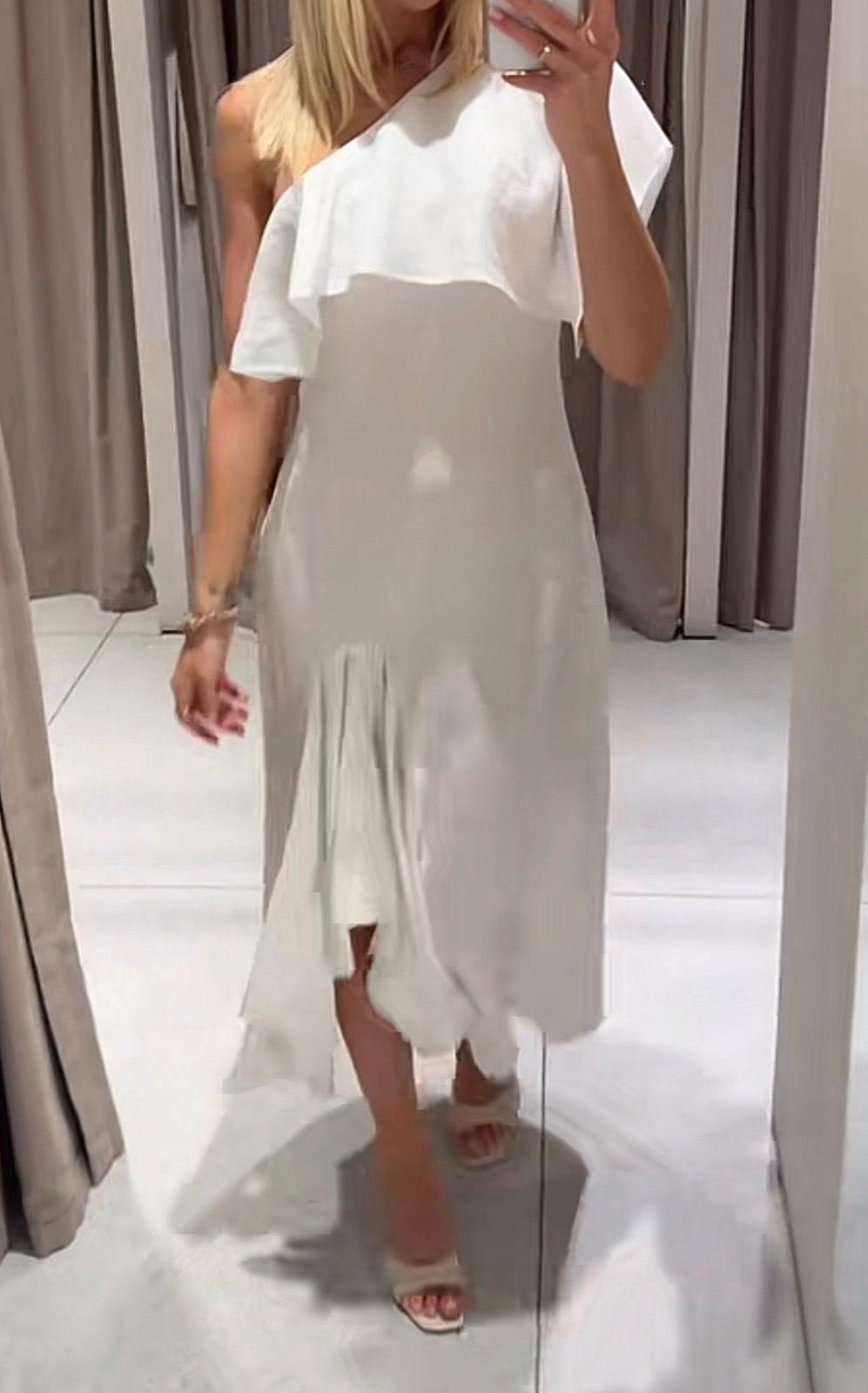 Woman taking a selfie in a changing room. She is wearing a white one-shoulder midi dress with a large frill, an asymmetric hem and heeled sandals.