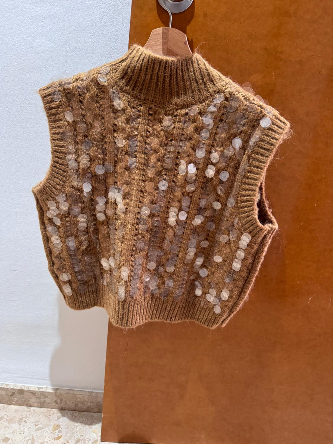 Brown knitted waistcoat with sequins hanging on a wooden hanger over a light brown door.